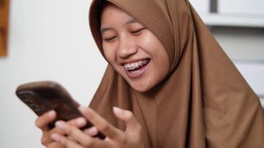 Genç Asyalı Müslüman kadın telefonuna baktığında gülümsüyor, mutlu bir yüz ifadesi, iyi haberler alıyor. 
