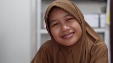 Genç Asyalı Müslüman kadın tesettüre girmiş, diş teli takmış kameraya bakıyor ve gülümsüyor. Mutlu ifade