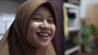 Genç Asyalı Müslüman kadın tesettüre girmiş, diş teli takmış kameraya bakıyor ve gülümsüyor. Mutlu ifade