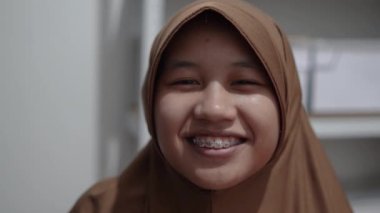 Genç Asyalı Müslüman kadın tesettüre girmiş, diş teli takmış kameraya bakıyor ve gülümsüyor. Mutlu ifade