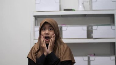 Genç Asyalı Müslüman kadın şok olmuş bir ifadeyle kameraya bakıyor, ağzı açık.