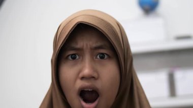 Genç Asyalı Müslüman kadın şok olmuş bir ifadeyle kameraya bakıyor, ağzı açık.