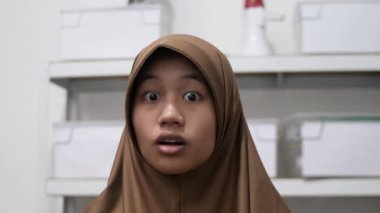 Genç Asyalı Müslüman kadın şok olmuş bir ifadeyle kameraya bakıyor, ağzı açık.