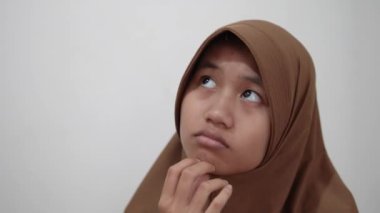 Asyalı genç bir müslüman kadın, eli çenesinde, düşünüyor ya da ciddi bir tefekkür ifadesi sergiliyor.