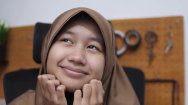 Genç, tatlı Asyalı Müslüman kız. Çenesini ellerinin üzerine koyup gülümsüyor ve merak ediyor. Mutlu bir ifade.