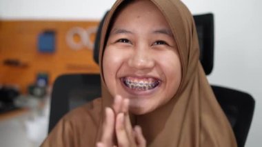 Genç Asyalı Müslüman kadın tesettüre girmiş, diş teli takmış kameraya bakıyor ve gülümsüyor. Mutlu ifade