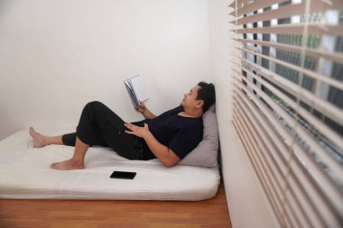 Yetişkin Asyalı adam uyumadan önce kitap okuyor, minimalist, yerde tek yataklı bir oda.