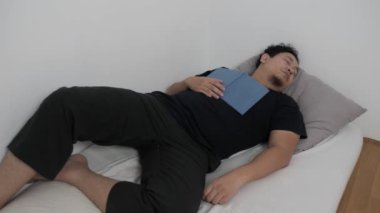 Yetişkin Asyalı bir adam kitap tutarken yatakta uyur. Yorgun, gevşemiş, kitap okuyan biri.