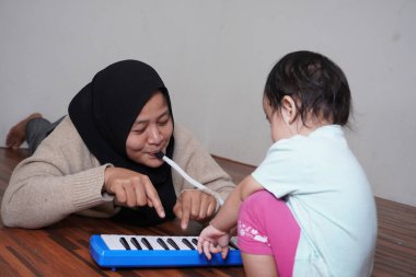 Asyalı Müslüman anne tesettüre girmiş, küçük kızıyla piyano çalıyor. Melodica bloworgan müzik enstrümanı.