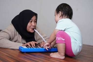 Asyalı Müslüman anne tesettüre girmiş, küçük kızıyla piyano çalıyor. Melodica bloworgan müzik enstrümanı.