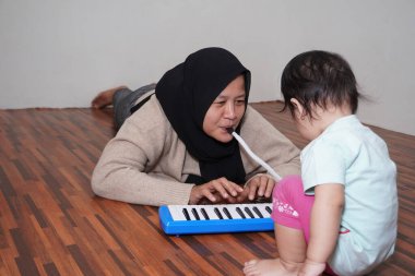 Asyalı Müslüman anne tesettüre girmiş, küçük kızıyla piyano çalıyor. Melodica bloworgan müzik enstrümanı.