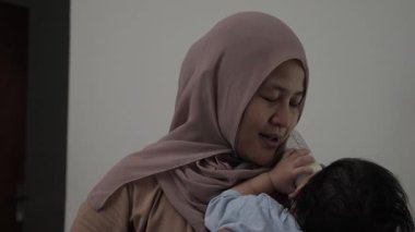 Asyalı Müslüman anne bebeğini kucağında bir şişeyle beslerken mutlu bir gülümsemeyle gülümsüyor.
