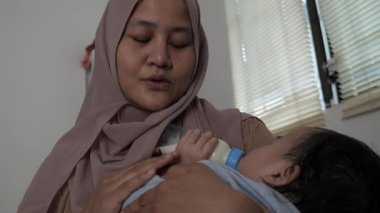 Asyalı Müslüman anne bebeğini kucağında bir şişeyle beslerken mutlu bir gülümsemeyle gülümsüyor.