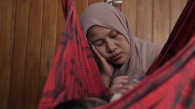 Asyalı Müslüman anne, bebeğini salıncakta sallarken yorgun ve uykulu görünüyor. Doğum sonrası depresyon