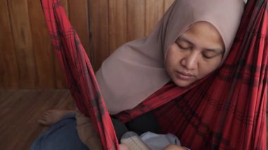Asyalı Müslüman anne, bebeğini salıncakta sallarken yorgun ve uykulu görünüyor. Doğum sonrası depresyon
