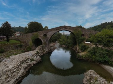 Nehir üzerindeki tarihi Ortaçağ Taş Köprüsü - Yeşil yemyeşil yeşillikler ve kayalık kıyılarla çevrili sakin ve yansıtıcı bir nehri geçen kalın sarmaşıklı tek kemerli taş köprü.