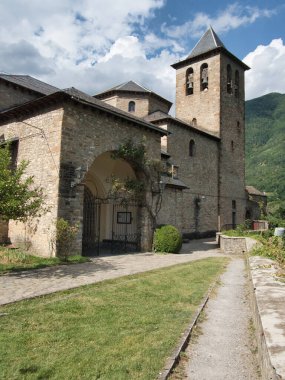 Tarihi Pyrenean Taş Kilisesi ve Kulesi - Uzun çan kulesi ve kemerli girişi olan ortaçağ taş kilisesi, yeşil dağ zeminine ve bulutlu gökyüzüne karşı çimenli bir yoldan görülüyor.