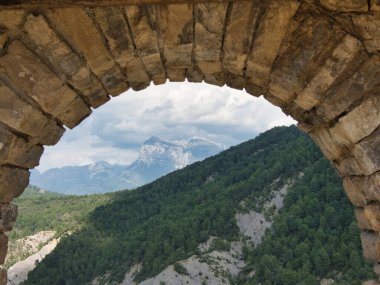 Stone Archway 'in çerçevelediği Mountain Vista - Çam ağaçlarıyla kaplı tepelerin ve kırsal, yıpranmış taş kemerlerin arasından görülen kayalık tepelerin güzel manzarası dramatik, bulutlu bir gökyüzünün altında.