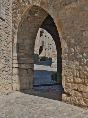 Village Caddesi 'ndeki Ortaçağ Taş Kemer Yolu, arka planda geleneksel taş evlerin bulunduğu tarihi, sessiz bir kasaba meydanına açılan güneşli, kemerli bir taş geçit..