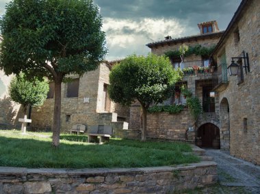 Sessiz Ortaçağ Köy Meydanı 'nda Taş Evleri - Bulutlu bir gökyüzünün altında geleneksel taştan binalarla çevrili iki kırpılmış ağaç ve taş bankları olan küçük çimenli meydan.