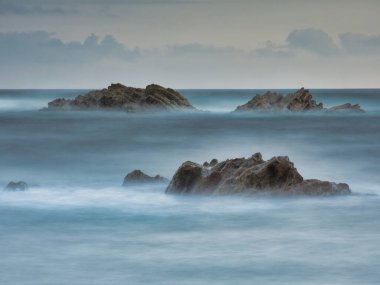 Moody Long Exposure Rocks in Blue Ocean Fog - Meydan formatında sivri uçlu deniz yığınlarının uzun pozlama fotoğrafı bulanık mavi ve turkuaz bir okyanus suyundan ortaya çıkıyor.