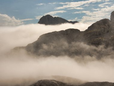 Bulutların tersine döndüğü engebeli tepenin üzerindeki engebeli tepe, kayalık bir tepenin üzerinde yükseltilmiş yürüyüş yolu, uzak, heybetli bir dağ sırasına bakan, mavi bir gökyüzüne bakan devasa bir bulut tepesi..