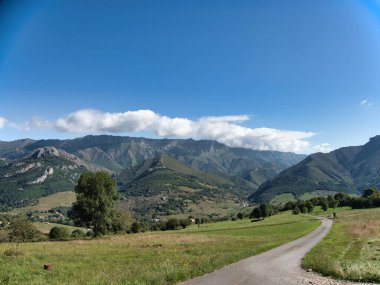 Asturias 'taki Dağ Vadisi' nde dolambaçlı yol - Parlak mavi bir gökyüzünün altında akan dağlarla yemyeşil çayırları kesen asfalt bir yolun manzarası.