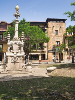 İspanyol Meydanı 'ndaki Süslü Tarihi Çeşme - Mavi gökyüzü altında geleneksel mimari ve yeşil ağaçlarla çevrili tarihi bir köy meydanındaki figürlerle detaylandırılmış taş çeşmesi.