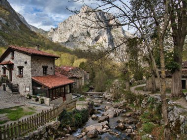River and Stone House ile Dağ Köyü - Kayalık nehir kenarındaki geleneksel taş evler, bulutlu bir gökyüzünün altındaki dramatik kireçtaşı dağları tarafından çerçevelenmiş.