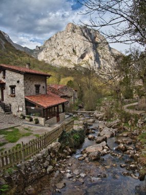River and Stone House ile Dağ Köyü - Kayalık nehir kenarındaki geleneksel taş evler, bulutlu bir gökyüzünün altındaki dramatik kireçtaşı dağları tarafından çerçevelenmiş.