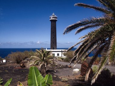 Palmiye ağaçları tarafından çerçevelenmiş volkanik siyah deniz feneri - uzun, siyah Faro de Fuencaliente (Fuencaliente Deniz Feneri) volkanik arazinin ve palmiye yapraklarıyla çerçevelenmiş Atlantik Okyanusu 'nun üzerinde yer alır..