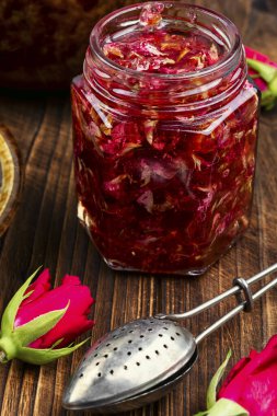 Gül reçelli kavanoz ve ahşap yüzeyde çiçek, gül yaprakları reçeli ve çay pişirme aletleri.