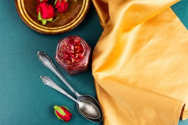 Gül reçelli kavanoz ve masada çiçek, gül yaprakları reçeli ve çay pişirme aletleri.