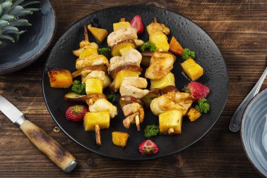Kızarmış tavuk parçaları ve bir tabakta ananas, ahşap yüzeyde meyve ve et şişleri..