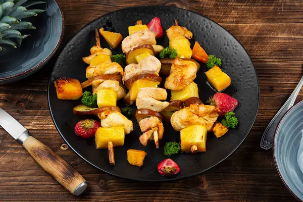 Kızarmış tavuk parçaları ve bir tabakta ananas, ahşap yüzeyde meyve ve et şişleri..