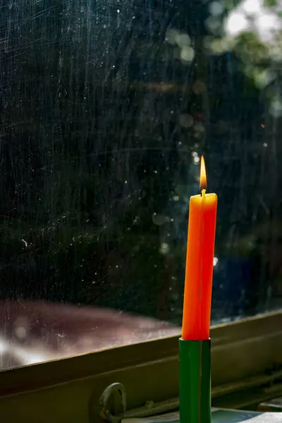A solitary orange candle casts a warm glow in a window, its flame flickering against a blurred outdoor background. The soft illumination creates a serene ambience in the evening darkness.