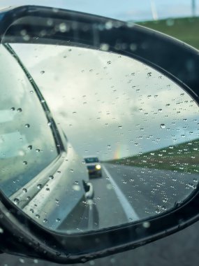 Yağmurlu bir gün sahnesi arabanın dikiz aynasında bulanık farları ve trafik yansımalarını gösteriyor. Yağmur damlaları camları serer, nemli, hareketli yola atmosferik bir bakış oluşturur..