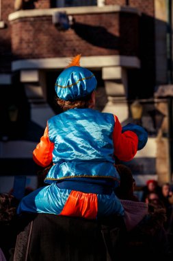 Dordrecht, Hollanda - 17 Kasım 2018: Pete kostümü giymiş, turuncu ve mavi giyinmiş genç bir çocuk, bayram kutlamaları sırasında merakla Aziz Nikolas 'ı bekleyen bir ebeveynin omuzlarında oturuyor..