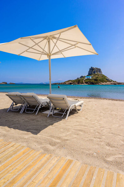Deck chair and umbrella on beautiful Agios Stefanos Beach in front of paradise Island Kastri- historical ruins and paradise scenery at coast of island Kos, Greece