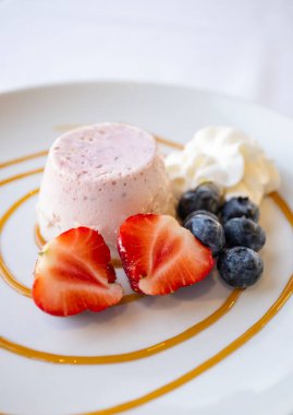 Panna Cotta, Mavi Böğürtlen, Çilek ve Açık Krema ile süslenmiş beyaz bir tabakta servis ediliyor.