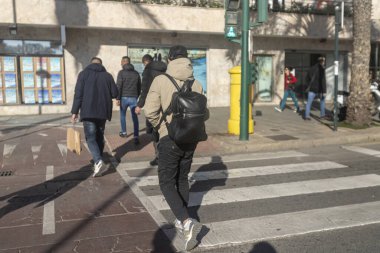 İnsanlar yaya geçidini geçerken sırt çantalı ve Faslı bir çocuğun sırt çantası ve şapkası takan genç bir çocuğun yakın çekimini yapıyorlar. Trafik ışığı da çok parlak.