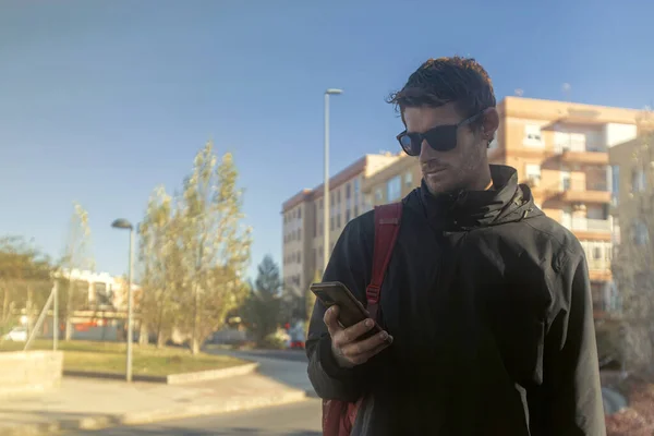 Telefonuna bakan genç bir adamın görüntüsü, şehirde ve bir sırt çantası takıyor ve güneş gözlüğü takıyor.