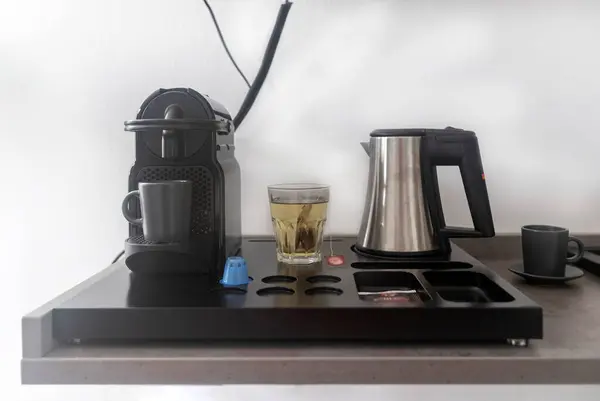 This photo features a breakfast set arranged on a table, including a capsule coffee machine, an electric kettle, a cup of coffee, coffee capsules, tea bags, and a glass of prepared chamomile tea. This setup offers a variety of options for a morning b
