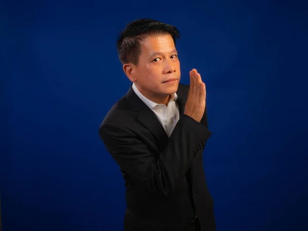 スタジオで青い背景に何かをささやくスーツを着て 魅力的で肯定的なプロのリーダーシップを見て 中高年アジアの自信に満ちた成功したビジネスマンの肖像画 — ストック写真