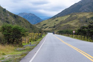 Soğuk bir iklimde ufukta kaybolan bir yolu ve yanında yeşil otlakları olan dağlık bir arazi.