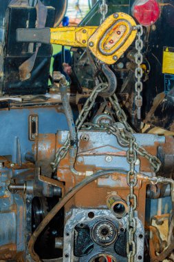 Eski, tozlu bir traktör motoru doğaçlama bir atölyede sarı bir makarayla zincirden sarkıyor..