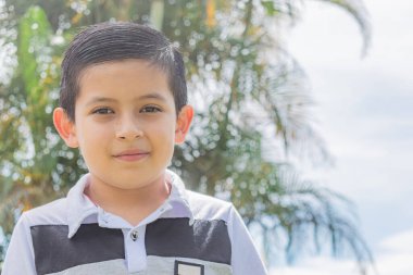 Güneşli bir günde, palmiye ağaçlarıyla dolu bir tarlanın ortasında kameraya bakan Latin bir çocuğun yüzüne yakın çekim.