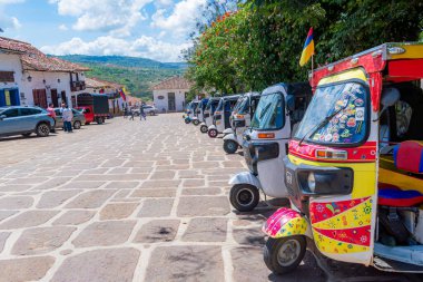 Barichara, Santander, Kolombiya, 11 Ekim 2024. Arka planda diğer tuk-tuk, turist ve araçlarla birlikte, ana meydana yakın ön planda canlı bir tuk-tuk duruyor. Bu renkli üç tekerlekli taksiler karakteristik ve kullanışlı bir araçtır.