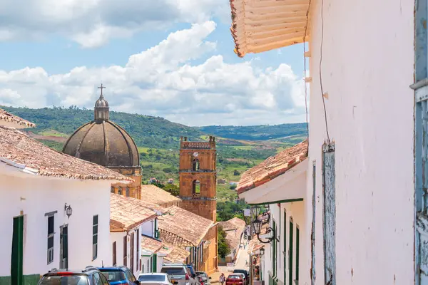 Barichara, Santander, Kolombiya, 11 Ekim 2024. Barichara 'daki ana katedralin dikey görüntüsü, kaldırım taşı sokağının sonunda belirgin bir şekilde duruyor. Yol boyunca birkaç araba park halindeyken, arka planda dağlar yükseliyor.