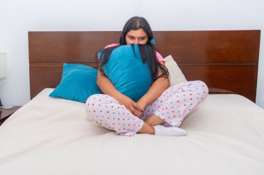 Genç Latin bir kadın yatakta oturuyor, yüzünde gözle görülür bir hüzün ile yastığa sıkıca sarılıyor. Kavram: Yalnızlık, üzüntü, duygusal mücadele.